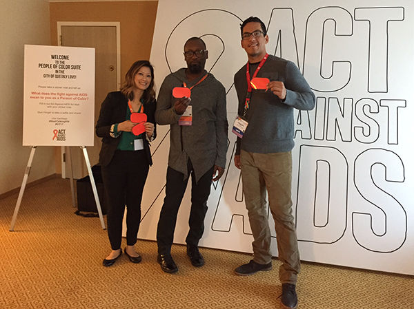 	Three members of the Act Against AIDS team standing in front of a large drawing of a ribbon