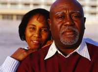 A man and a woman standing.  The middle-aged man is in the foreground, looking into camera.  The woman is standing behind him with her hands on his shoulder, smiling and also looking into the camera.