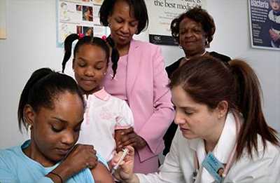 A nurse in the process of administering an intramuscular vaccination into a girl’s left arm whose sister, mother, and grandmother watched from the background