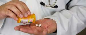 Male physician in white coat dispensing to white round tablets from a medicine bottle. He's wearing a stethoscope. 