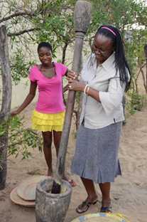 Naemi Shoopala, CDC-Namibia Field Office Coordinator, takes a break from a site visit to try her hand at grinding the grains used to make porridge in a small settlement outside Rundu, while talking to a teenage girl about HIV prevention