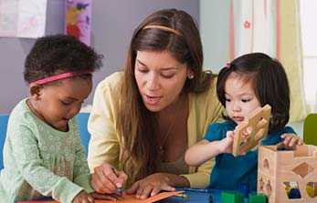 	A teacher is helping a toddler draw while another toddler is stacking blocks.