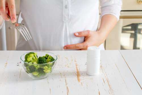 Pregnant female choosing between broccoli in glass bowl and vitamins.