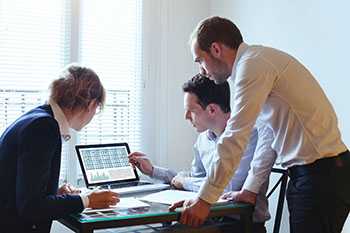 two me and a woman gathered around a lap top examining information on the screen