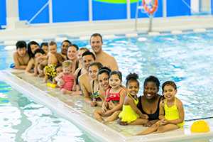 An image of a long float in a pool with children sitting in a line. In the water next to the children stands two instructors.