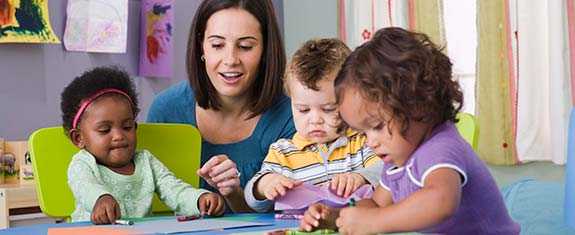 A woman supervising three young children working on art projects. 