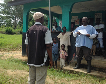 	Fig 1.c. This image shows the clinic’s waiting area behind the OIC where multiple patients wait for care. Allowing uncontrolled access by patients with suspected EVD into a common waiting area poses risks to patients and HCWs. Here the OIC practices telling a suspected EVD patient’s father to bring his son to another entrance where suspect and probable EVD cases can be isolated and cared for while awaiting transfer to an ETU. (Photo credit: Miwako Kobayashi, CDC EISO. All persons provided verbal consent to being photographed.)